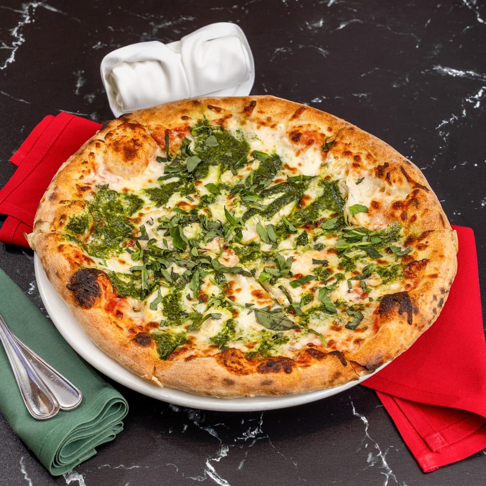 A wood-fired pesto pizza with melted cheese and fresh basil on a white plate with red and green napkins.