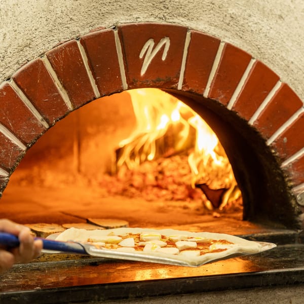 A pizza with cheese and sauce on a peel, entering a wood-fired oven with flames.