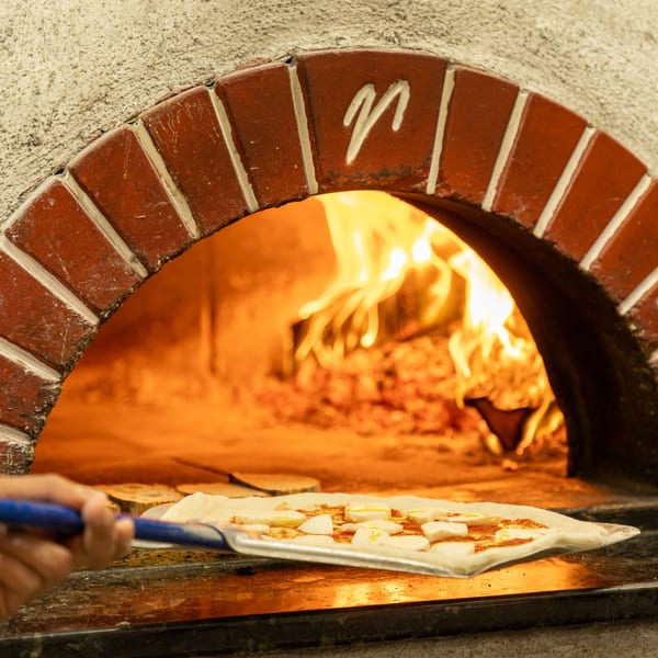 A pizza with mozzarella and sauce on a peel, ready to enter a brick wood-fired oven.