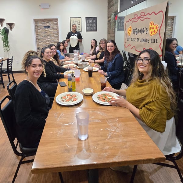 A group of women making pizzas at a long wooden table, with a man holding pizza dough.