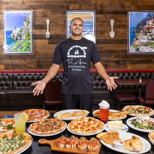A man in a Ti Amo Wood Fired Pizza shirt stands behind a table laden with pizzas and other dishes.