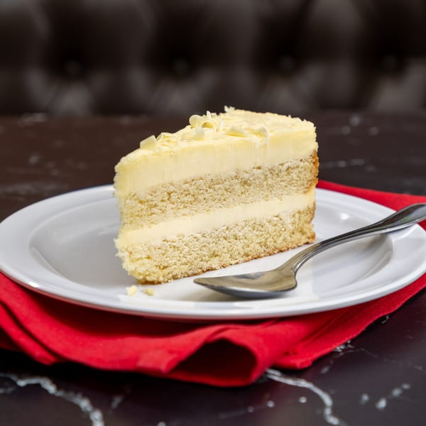 A slice of vanilla cake with white frosting and shavings on a white plate with a spoon.