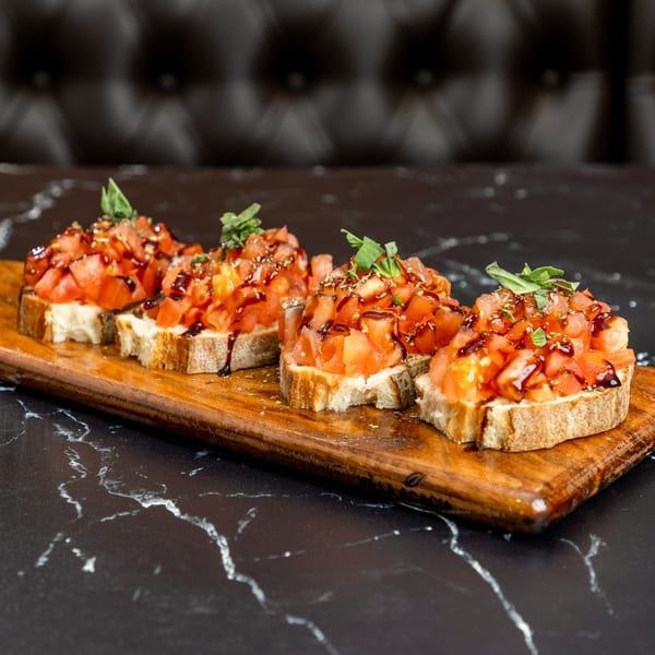 Four bruschetta appetizers with diced tomatoes, basil, and balsamic glaze on a wooden board.