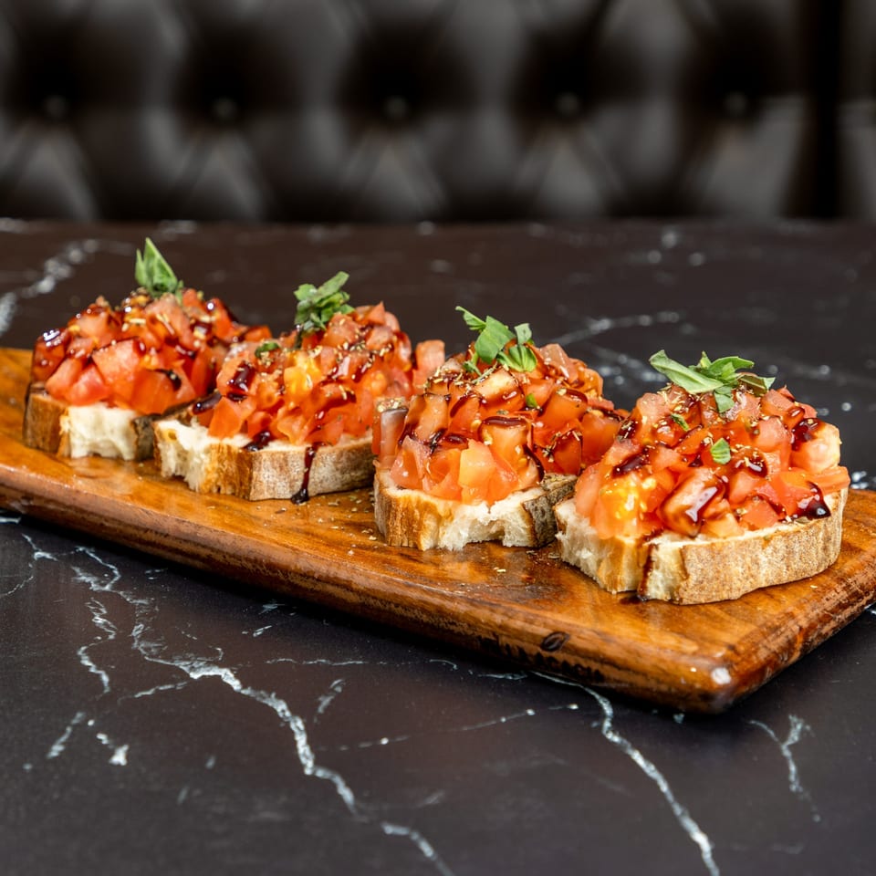 Four bruschetta appetizers with diced tomatoes, basil, and balsamic glaze on a wooden board.