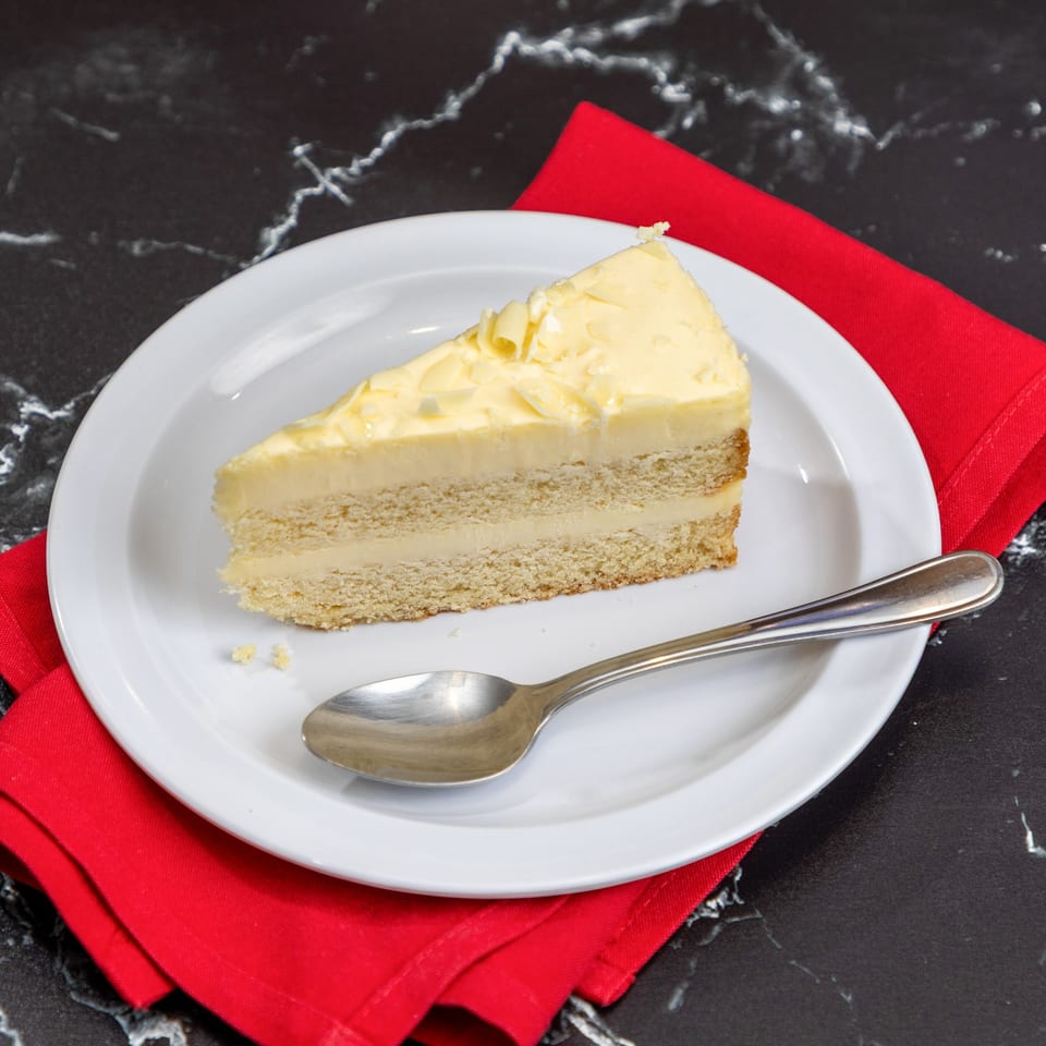 A slice of white chocolate cake with shavings on a white plate with a spoon and red napkin.