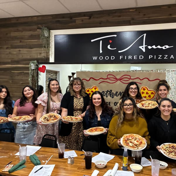 Ten women smiling, holding individual pizzas, standing in front of the Ti Amo Wood Fired Pizza sign.