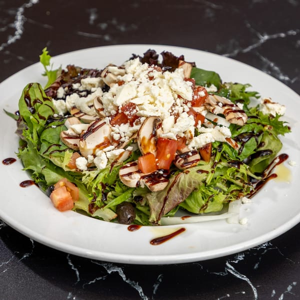 Fresh green salad with mushrooms, tomatoes, feta cheese, and balsamic glaze on a white plate.