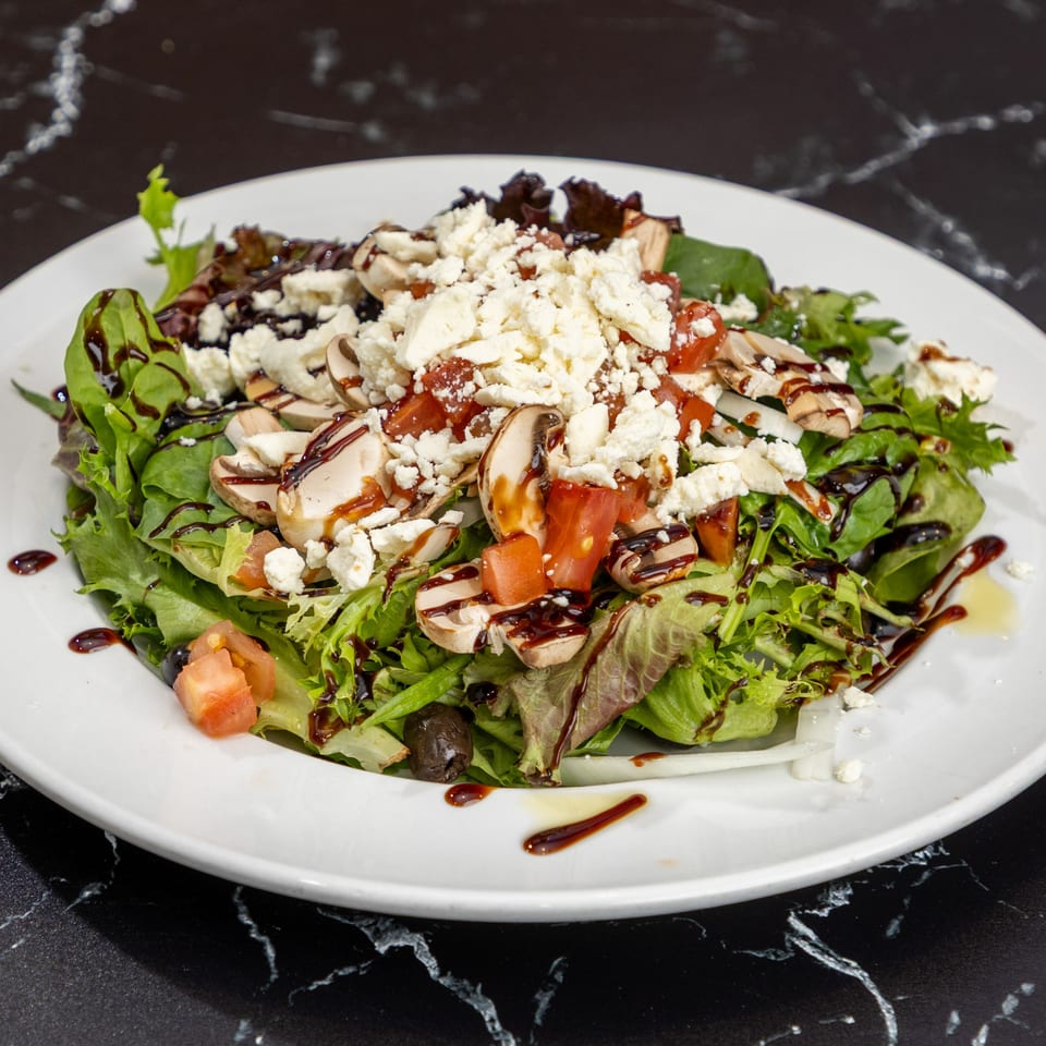 Fresh green salad with mushrooms, tomatoes, feta cheese, and balsamic glaze on a white plate.