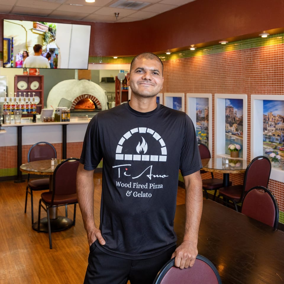A man in a Ti Amo Wood Fired Pizza shirt smiles in his restaurant with a pizza oven behind him.