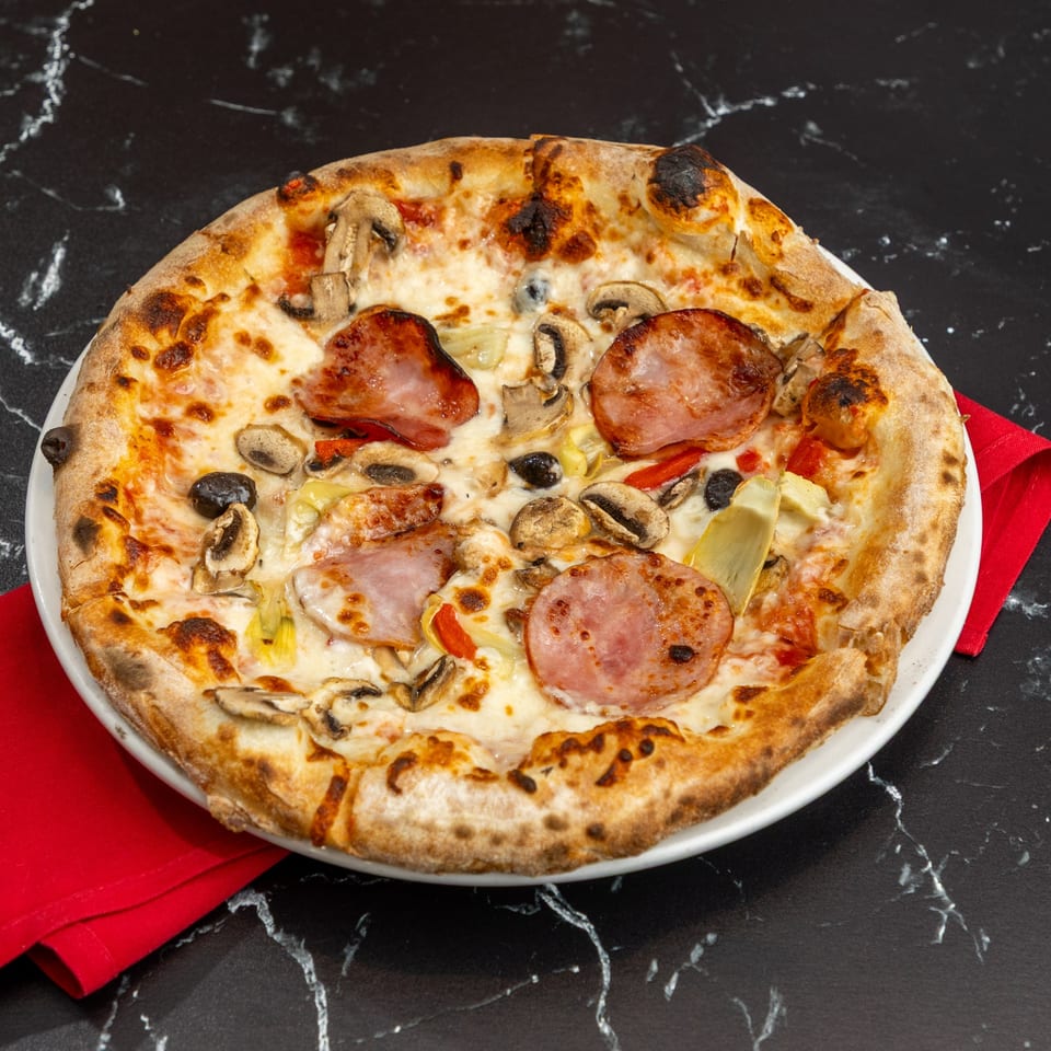 Ti Amo Wood Fired Pizza with ham, mushrooms, olives, and artichoke hearts on a black marble surface.