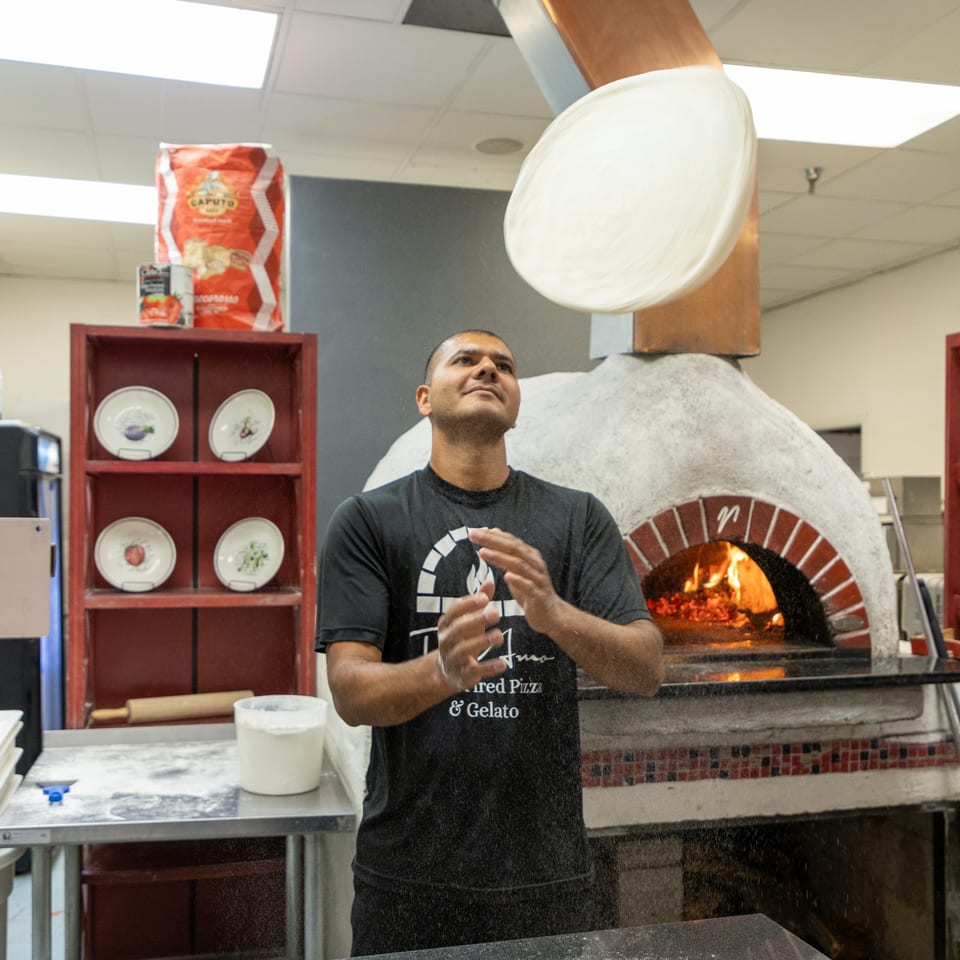 A Ti Amo chef expertly tosses pizza dough high above a wood-fired oven, flour dusting the air.